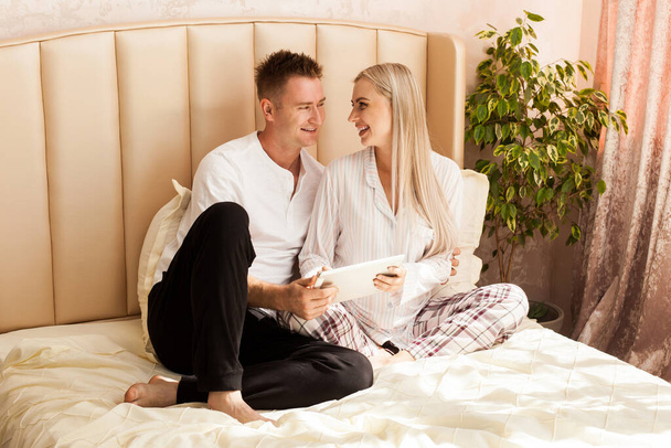 A pregnant woman and a man are lying on the bed at home. They are looking at something with interest on their white tablet, choosing a name for the baby - Fotografie, Obrázek