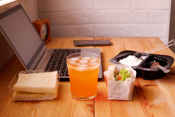 Φάτε σάντουιτς και το γεύμα πλαστικό κουτί με χυμό πορτοκαλιού. Κατά τη διάρκεια της εργασίας με φορητό υπολογιστή. Δούλεψε και φάε ταυτόχρονα. Εργασία στο σπίτι. - Φωτογραφία, εικόνα