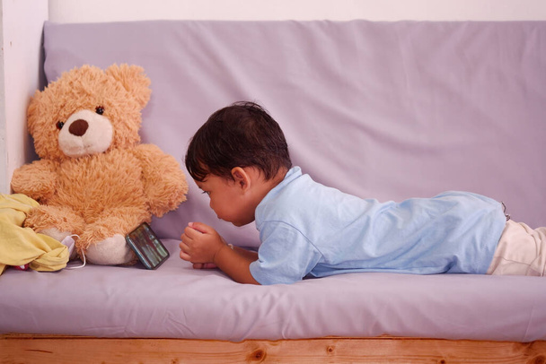 Asiatischer Junge ca. 2 Jahre alt, beobachtet und spielt Handy sitzt auf dem Sofa im Wohnzimmer. Den Teddybär ignorieren - Foto, Bild
