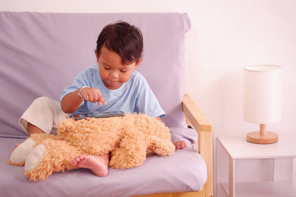 Süße Asiatin ca. 2 Jahre alt, beobachtet und spielt Handy sitzt auf dem Sofa im Wohnzimmer. Den Teddybär ignorieren - Foto, Bild