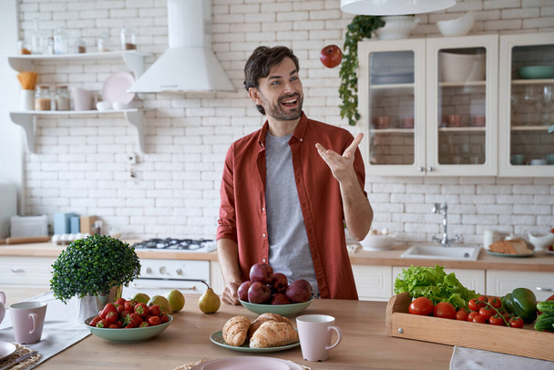Her sabah elma yiyorum. Genç mutlu sakallı adam, gündelik kıyafetlerle kırmızı elma kusuyor ve modern mutfakta dururken gülümsüyor. - Fotoğraf, Görsel