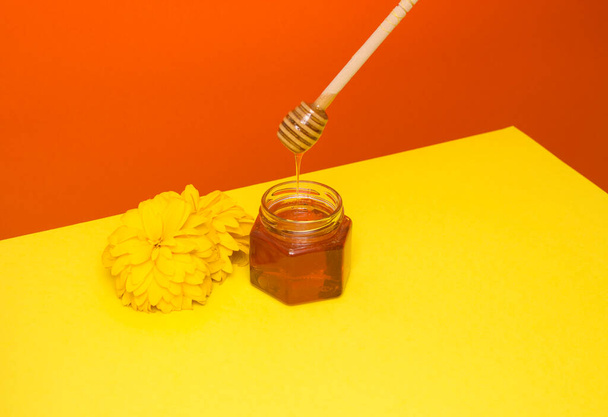 Včelí med kape z dřevěné špachtle do skleněné nádoby. Květinové, žlutočervené pozadí. Užitečné zemědělské produkty. - Fotografie, Obrázek