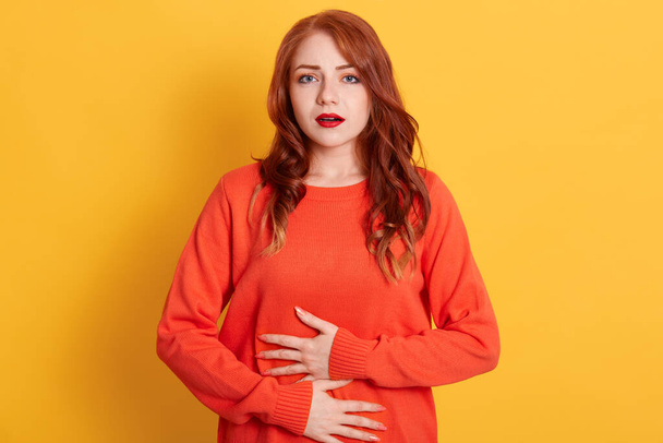Woman with stomachache, having food poisoning, posing isolated over yellow background, looks at camera, lady has red wavy hair wearing casual orange sweater. - Foto, afbeelding