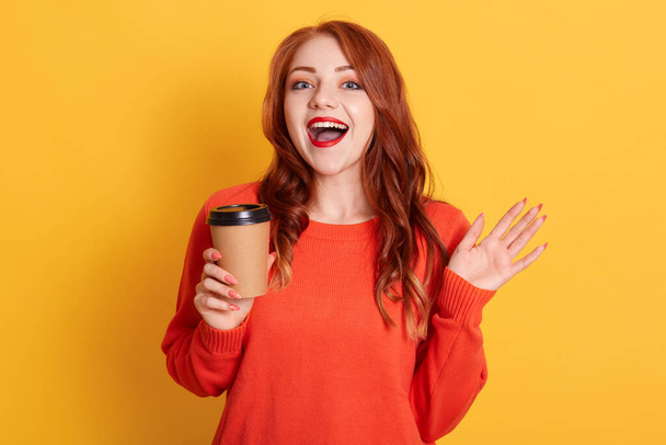 Pretty red haired female prefers coffee take away, holds disposable cup with aromatic hot drink, looking at camera with glad expression and toothy smile, wears orange sweater. - Fotoğraf, Görsel