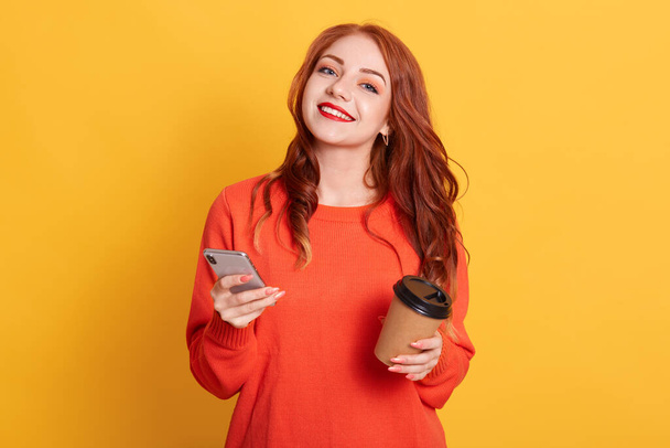 Pleased European woman dressed in casual orange sweater, poses over yellow background, looks smiling at camera, holding smart phone and coffee to go. - Foto, Imagen
