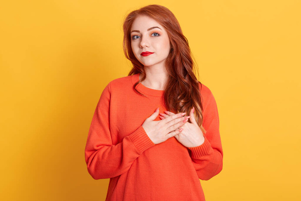 Lovely female with friendly look, keep hands on chest, expresses positive and good attitude, has red wavy hair, feels gratitude to her mother, posing isolated over yellow background. - Foto, afbeelding
