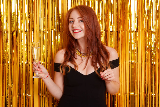 Portrait of young attractive woman wearing black dress drinking champagne and celebrating Christmas or birthday on wall with tinsel decoration, smiling girl. - Фото, изображение