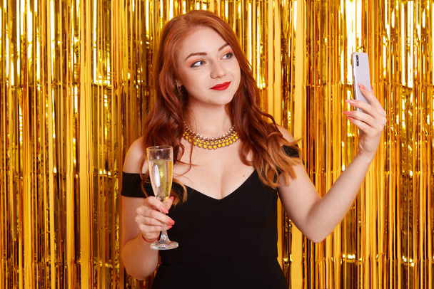 Red haired female wearing black dress and necklace making selfie photo via her smart phone, holding glass of wine, posing isolated over wall decorated wit golden tinsel. - Zdjęcie, obraz
