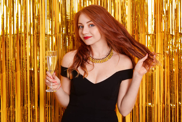Happiness woman holding glass of champagne, touching her red beautiful hair, posing against golden tinsel on background, lady in black dress on party. - Φωτογραφία, εικόνα