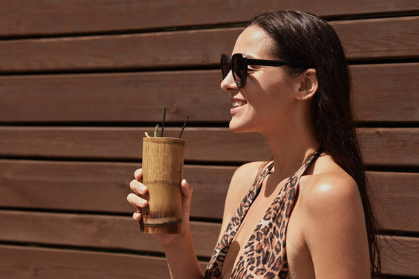 Happy woman enjoying exotic drink in bar, looking smiling in distance, wearing black sunglasses and swimming suit, holding non alcoholic beverage against brown wooden wall. - Photo, Image