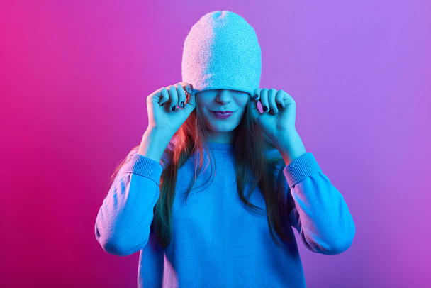 Attractive woman pulling hat over her eyes wearing casual sweater posing isolated over pink neon background, young lady with long hair having fun, - Foto, imagen