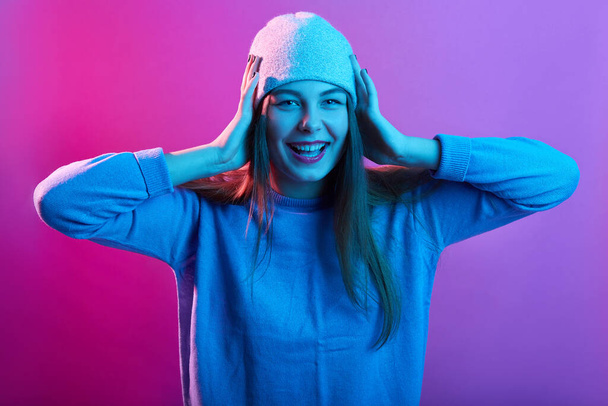 Portrait of young female with long hair looking in excitement at camera, excited girl in casual sweater and cap. Human face expressions posing against pink neon wall. - Fotó, kép