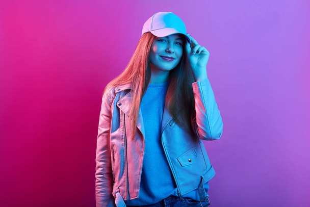 Fashion portrait of confident woman standing against pink neon wall and touching visor of her cap, wearing leather jacket, looking at camera, has pleasant appearance. - Фото, изображение