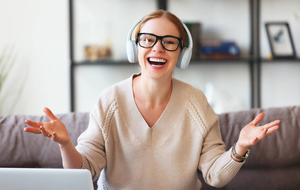 Happy adult female in glasses and headphones gesticulating and laughing while sitting on sofa at home during a video cha - Fotoğraf, Görsel
