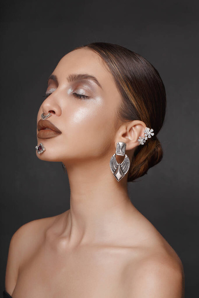 Profile portrait of a girl with makeup and jewelry applied to the face, with bare shoulders, with closed eyes, posing with attitude, on a gray background. Vertical view. - Фото, зображення