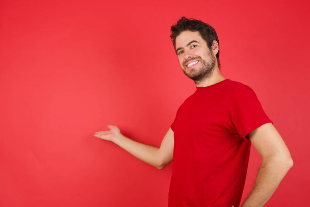 Νεαρός όμορφος μελαχρινός άντρας με κόκκινη μπλούζα που νιώθει χαρούμενος και χαρούμενος, χαμογελαστός και σε καλωσορίζει, προσκαλώντας σε με μια φιλική χειρονομία  - Φωτογραφία, εικόνα