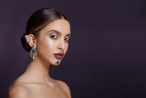 Profile portrait of a girl with splendid makeup and hair arranged, pose in the studio in profile with bare shoulders, jewelry with ears, neck, nose, chin, over dark background, copy space. - Photo, Image