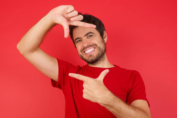 孤立した赤い背景にTシャツを着た若いハンサムな白人男性が手で指のフレームを作ります。創造性と写真の概念. - 写真・画像