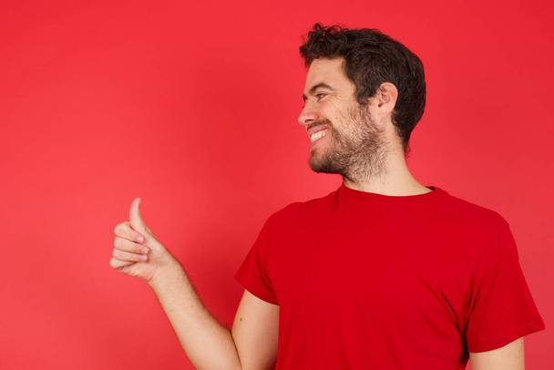 Junger gutaussehender kaukasischer Mann mit T-Shirt vor isoliertem roten Hintergrund, der stolz lächelt und Daumen nach oben gestikuliert. Gute Arbeit! - Foto, Bild