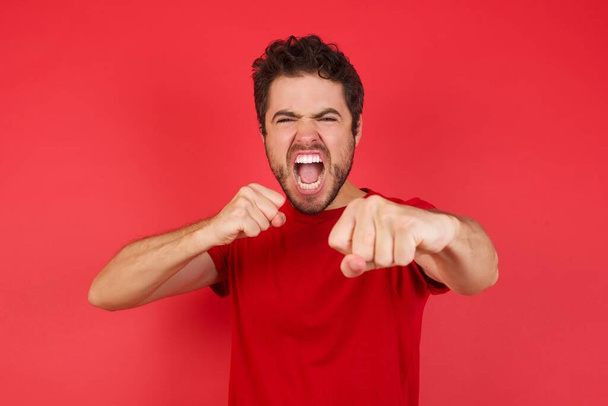 Portret van een sterke en vastberaden Jonge knappe blanke man die een t-shirt draagt over een geïsoleerde rode achtergrond die lucht met vuist slaat en vol vertrouwen naar de camera kijkt, mannelijke strijd, vechtlust.  - Foto, afbeelding