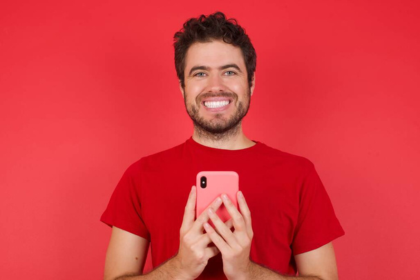 Молодой красивый кавказский мужчина в футболке на изолированном красном фоне пользуется дистанционным общением, пользуется мобильным телефоном, пользуется быстрым безлимитным интернетом, имеет приятную улыбку, одет в красную рубашку, делает покупки в Интернете,  - Фото, изображение