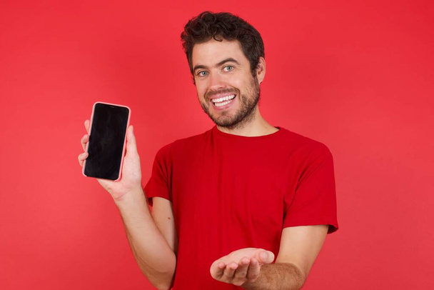 Jonge knappe blanke man draagt t-shirt over geïsoleerde rode achtergrond met een mobieltje. presentatie smartphone  - Foto, afbeelding