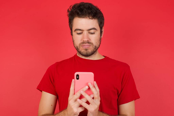 Retrato de hombre caucásico joven guapo emocionado usando camiseta sobre fondo rojo aislado guiño y ojo mantenga el uso del teléfono inteligente leer noticias de redes sociales  - Foto, imagen