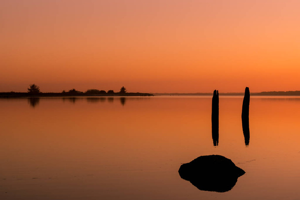Very orange sunset with pilings and logs reflect in glassy river surface - Photo, Image