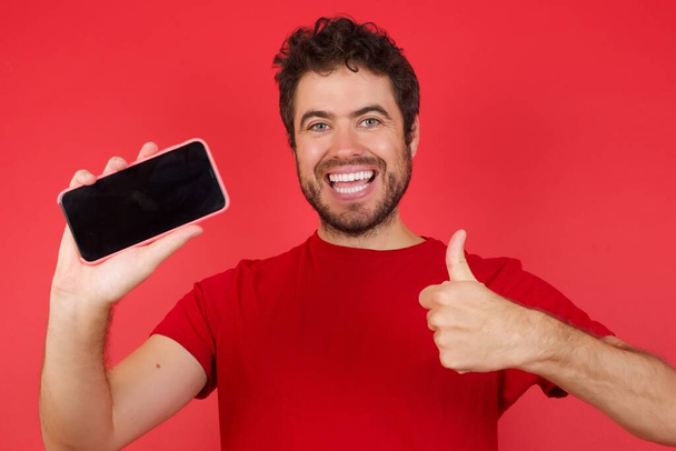 Retrato Joven hombre caucásico guapo usando camiseta sobre fondo rojo aislado, sosteniendo en las manos la celda que muestra dando pulgar pantalla negra hacia arriba  - Foto, Imagen
