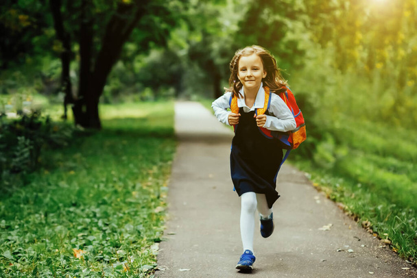 A girl in a school uniform and a backpack runs along the road to the park, the concept of the beginning of the school year - Valokuva, kuva