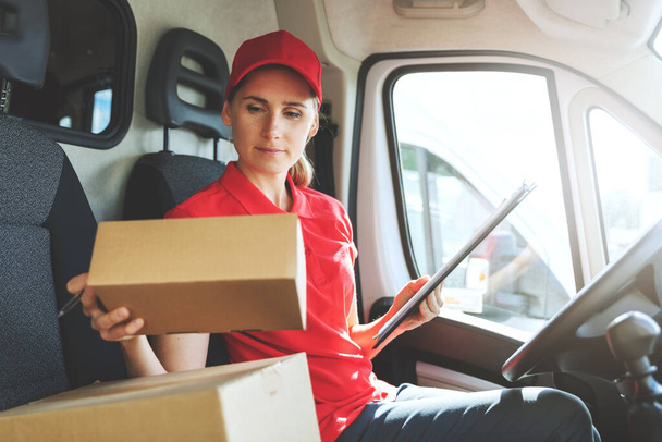 работница службы доставки женщин в красной форме сидит в фургоне и работает с пакетами - Фото, изображение