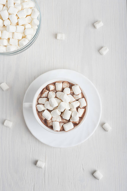 View from above on cup full of sweet homemade cocoa drink usually prepared at cold winter vacations served on plate with bowl of marshmallow on white wooden background. Vertical orientation image - Foto, imagen
