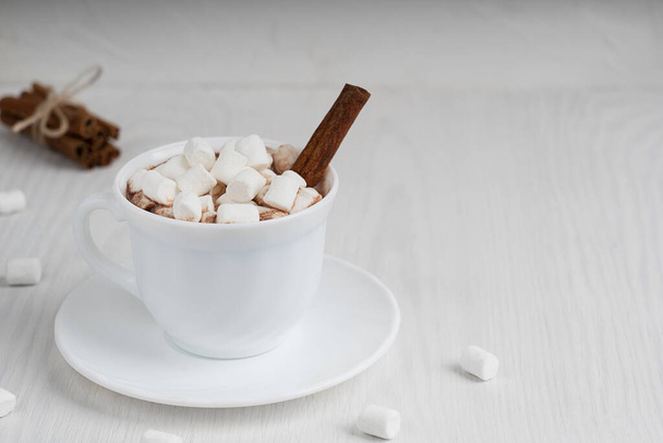 Cup full of hot homemade cacao drink usually prepared at cold winter and autumn seasons served on plate with marshmallows and cinnamon on white wooden background. Image with copy space, horizontal - Fotografie, Obrázek
