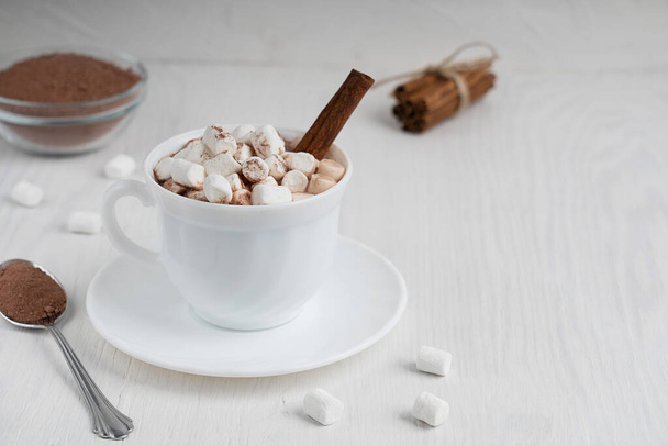 Cup full of hot homemade cacao drink usually prepared at cold winter days served on plate with marshmallows, cinnamon and spoon on white wooden background at kitchen. Image with copy space, horizontal - Foto, Bild