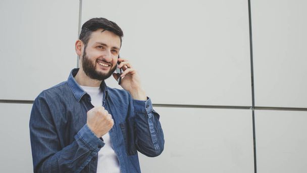 Happy young bearded man talking on mobile phone and delighted clench fist victory triumph gesture, hearing good news in conversation standing on grey wall background. Celebrate success, achieve goal. - Foto, Imagen