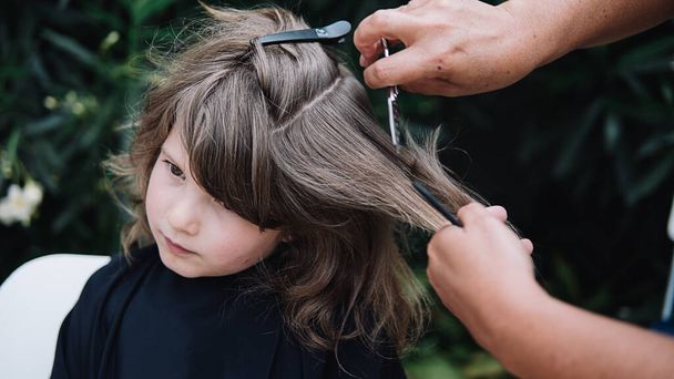 Készletkép fiatal lányról, akinek levágatja a haját egy nő a kertben. A fodrász ollót használ.. - Fotó, kép