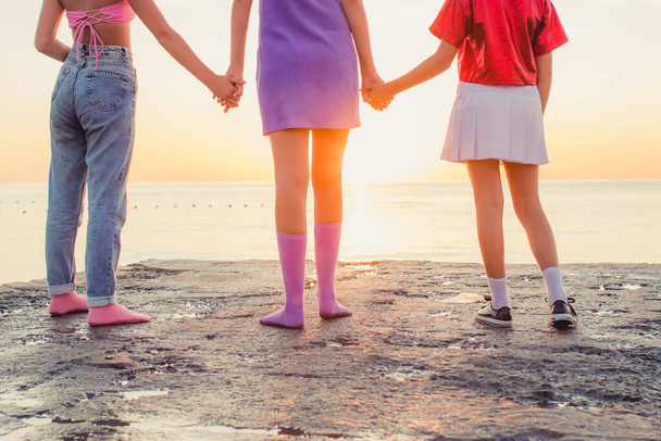 Вид ззаду трьох молодих друзів, що стоять на пляжі, тримаючись за руки і дивлячись на море на сході сонця
. - Фото, зображення