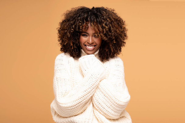 Happy beautiful african girl with afro hairstyle posing in cozy sweater on beige studio background. - Photo, Image