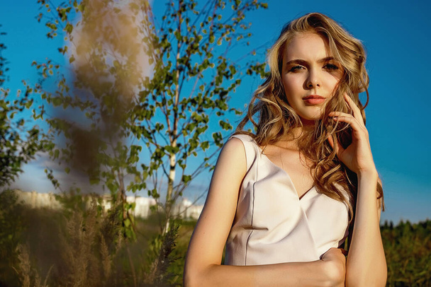 Portrait of a blonde young girl in a dress that sits in a field with tall grass against a background of blue sky and a tree. On a sunny day at sunset at golden hour in summer - Photo, image