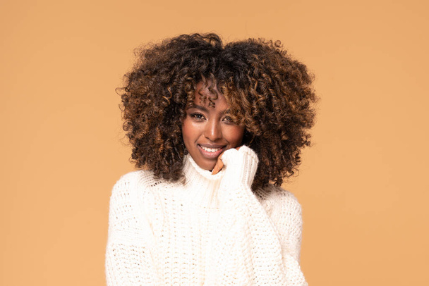 Happy beautiful african girl with afro hairstyle posing in cozy sweater on beige studio background. - Foto, immagini