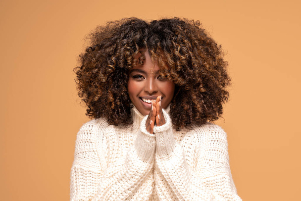 Happy beautiful african girl with afro hairstyle posing in cozy sweater on beige studio background. - 写真・画像