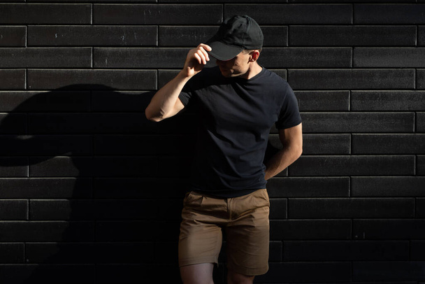 Mężczyzna w czarnej koszulce i czapce bejsbolowej pozuje na czarną ścianę - Zdjęcie, obraz