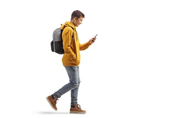 Full length profile shot of a male teenage student walking and looking at a mobile phone isolated on white background - Photo, Image