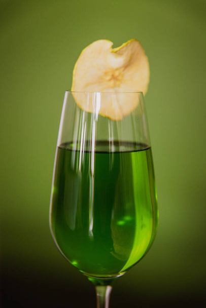 A close-up of a green cocktail in a flute glass garnished with a dehydrated apple slice on green background - Foto, imagen