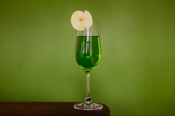 A green cocktail in a flute glass garnished with a dehydrated apple slice on green background - Foto, Imagen