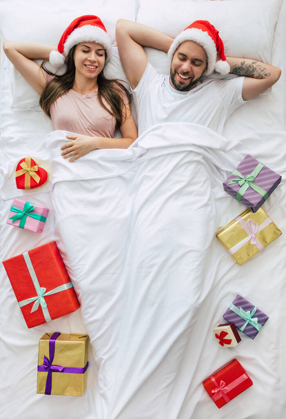 Top view full-length photo of a beautiful young happy couple in Santa hats with many Christmas gift boxes and dreaming while sleeping. Above photo. Merry Christmas and happy new year - Foto, afbeelding