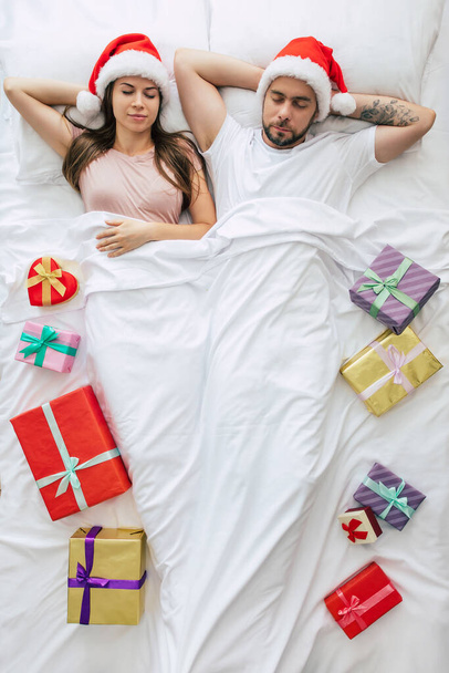 Зверху - повноформатне фото красивої молодої щасливої пари в Санта-Капелюшках з великою кількістю різдвяних подарунків і снами під час сну. На фото зверху. Веселого Різдва і щасливого нового року - Фото, зображення