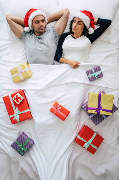 Top view full-length photo of a beautiful young happy couple in Santa hats with many Christmas gift boxes and dreaming while sleeping. Above photo. Merry Christmas and happy new year - Фото, изображение