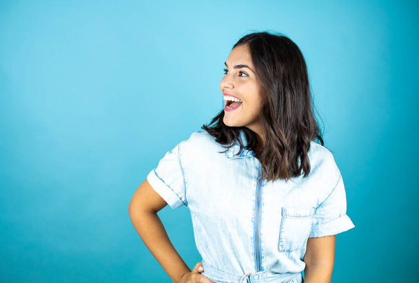 Jonge mooie vrouw draagt een spijkerbroek over geïsoleerde blauwe achtergrond glimlachend en kijkend naar de lege ruimte aan de zijkant. - Foto, afbeelding