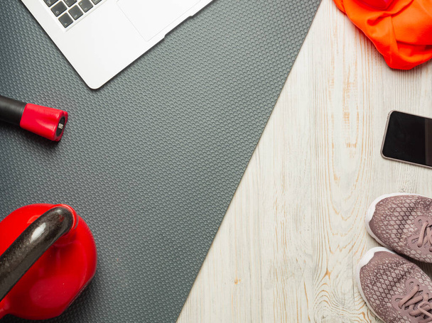 Fitness concept - sports and fitness at home (laptop, bodybar, red kettlebell, gray mat, wooden light floor, sneakers, phone, orange towel), flat lay, top down, copy space, horizontal orientation - Photo, image